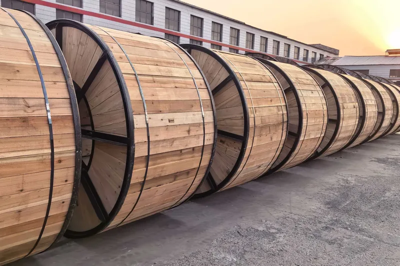 A long row of empty wooden cable drums with dark steel bands, lined up on the concrete yard outside the factory, with workshop buildings and the setting sun behind them